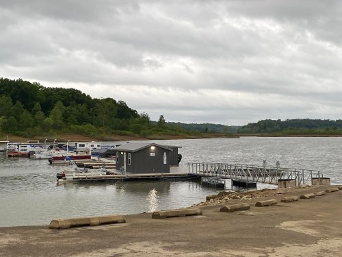 A gray dock with a small building on a calm lake, surrounded by green trees and cloudy skies. Several boats are moored nearby.