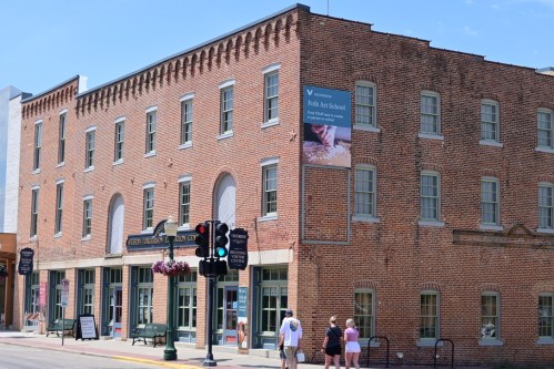 Historic brick building with large windows, a banner for a folk art school, and pedestrians at a traffic light.