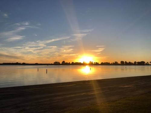 A serene sunset over a calm lake, with reflections on the water and silhouettes of trees in the background.