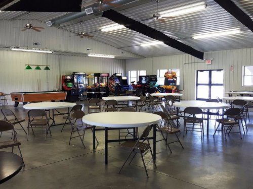 A spacious indoor area with round tables and chairs, featuring arcade games in the background.