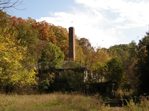 An abandoned house surrounded by trees with autumn foliage and a tall chimney against a cloudy sky.