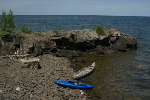 Two kayaks, one blue and one gray, rest on a rocky shore by a calm lake under a clear blue sky.