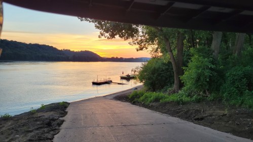 Sunset over a calm river, with a boat dock and lush greenery along the shore.