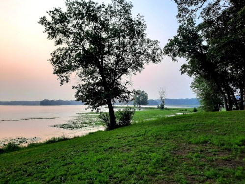 A serene lakeside view at sunset, featuring trees and lush green grass along the water's edge.