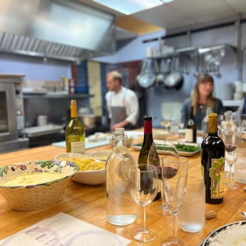 A busy kitchen with a chef in the background, wine bottles, glasses, and plates of food on a wooden table.