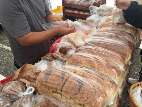 A person handling loaves of bread wrapped in plastic at a market stall, surrounded by more baked goods.
