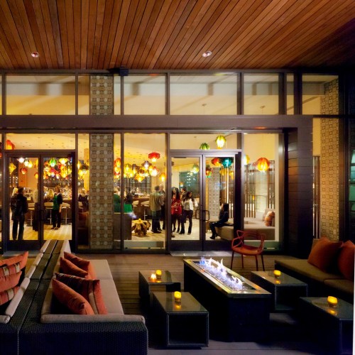 A cozy lounge area with colorful pendant lights, modern seating, and a fire pit, overlooking a lively indoor space.