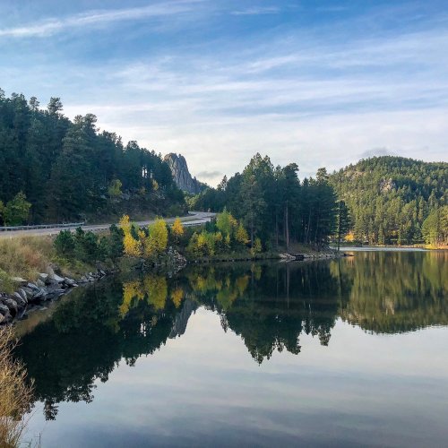 A serene lake reflects trees and mountains under a clear sky, with autumn colors along the shore.