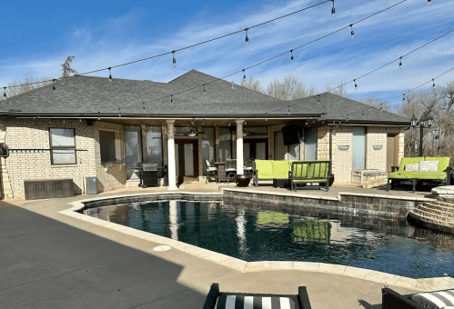 A backyard with a pool, patio furniture, and string lights, set against a clear blue sky and a brick house.