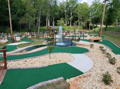 A mini-golf course with a fountain, surrounded by greenery and pathways, featuring players enjoying the game.
