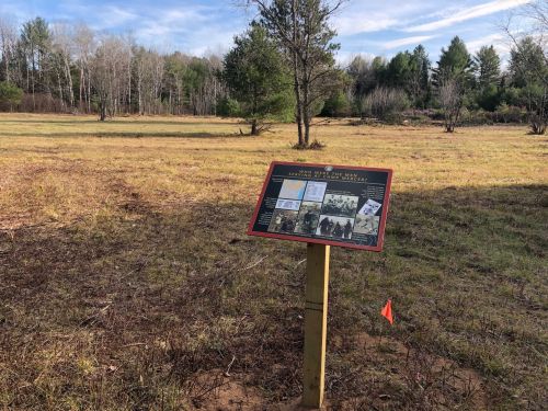 A sign on a post in a grassy field, surrounded by trees, displaying historical information and photos.