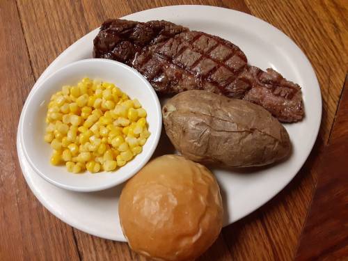 A plate with grilled steak, a baked potato, corn, and a dinner roll on a wooden table.