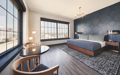 Modern hotel room featuring a king bed, stylish decor, a desk, and large windows with city views.