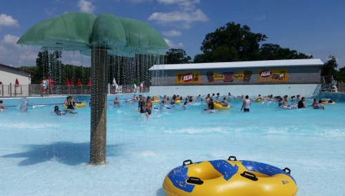 A water park scene with a palm tree fountain, people swimming, and colorful inner tubes in a blue pool under a sunny sky.