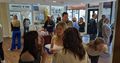 A busy indoor event with people socializing, a table with displays, and decorations celebrating MHACC @ 100.