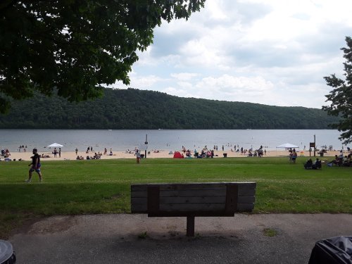 A scenic view of a beach with people enjoying the water, surrounded by green hills and trees under a cloudy sky.