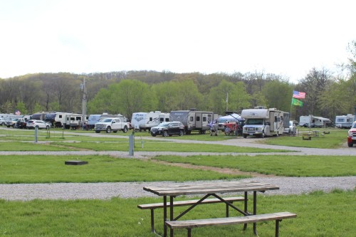 A campground with several RVs parked, green grass, a picnic table, and trees in the background under a cloudy sky.
