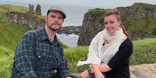 A man and woman sit on a rocky ledge overlooking a scenic coastal landscape with greenery and ocean in the background.