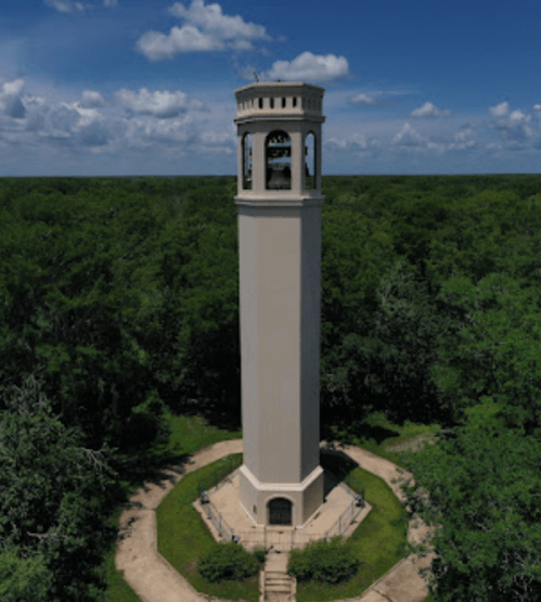 A tall, white tower surrounded by lush green trees under a blue sky with scattered clouds.