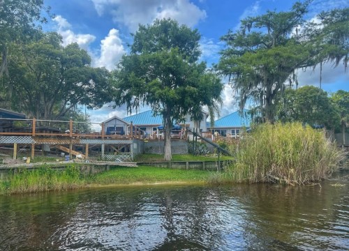 A scenic view of a wooden deck by a river, with a house and lush greenery under a blue sky with clouds.