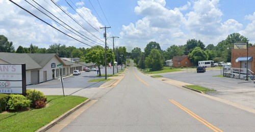 A quiet street lined with trees and buildings under a blue sky with fluffy clouds. Power lines run along the road.