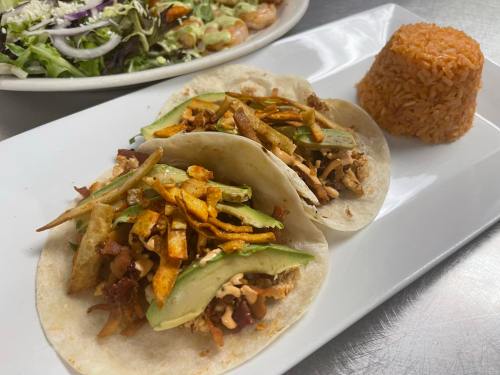 Two tacos filled with meat, avocado, and crispy strips, served with a side of rice and a fresh salad in the background.