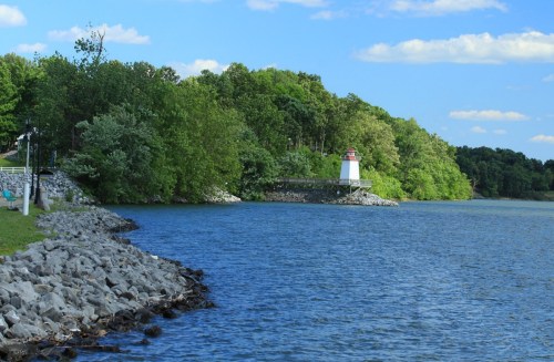 A serene lakeside view featuring a lighthouse surrounded by lush greenery and a rocky shoreline under a blue sky.