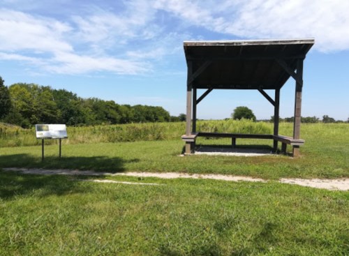 A wooden shelter with a bench beside a grassy field and a sign, under a blue sky with scattered clouds.