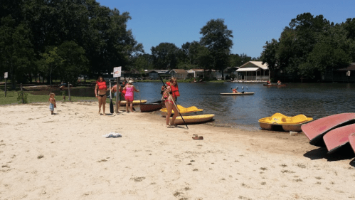 A sandy beach with people in life jackets near yellow paddle boats, while others kayak on a calm lake.