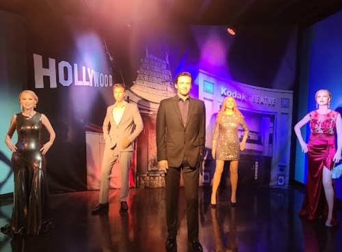 Five lifelike wax figures in glamorous outfits stand in front of a Hollywood backdrop, featuring the Kodak Theatre.