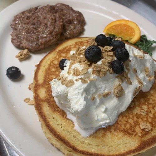 A plate with a large pancake topped with whipped cream, blueberries, and granola, alongside two sausage patties and an orange slice.