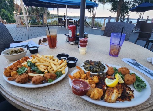 Two plates of seafood with fries, hushpuppies, and greens, set on a table with drinks and a scenic outdoor view.