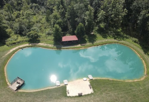 Aerial view of a serene pond surrounded by trees, with a small cabin and a sandy area by the water's edge.