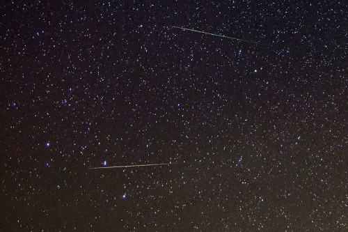 A starry night sky filled with numerous stars and two bright meteor trails streaking across the scene.