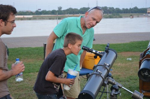 A man and a boy observe through a telescope by a river, with another person nearby holding a water bottle.
