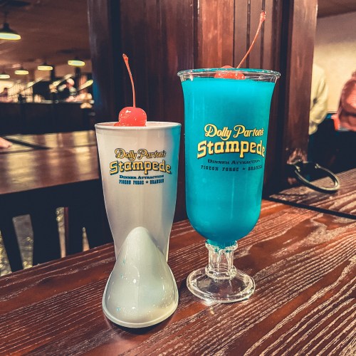 Two colorful drinks with cherries on top, featuring the logo of Dolly Parton's Stampede, on a wooden table.