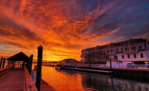 A vibrant sunset paints the sky in orange and purple over a calm waterfront with a wooden pier and buildings.