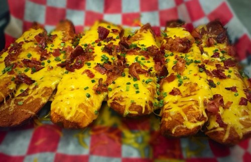 A close-up of crispy potato wedges topped with melted cheese, bacon bits, and green onions on a red checkered paper.