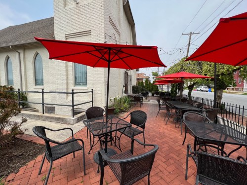 Outdoor patio with black tables and chairs, featuring red umbrellas, beside a white brick building.