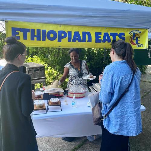 A vendor serves food at an Ethiopian eats stall, with two customers browsing the offerings under a tent.
