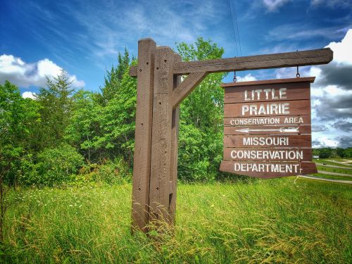 Wooden sign for Little Prairie Conservation Area, surrounded by greenery and blue skies, indicating Missouri Conservation Department.