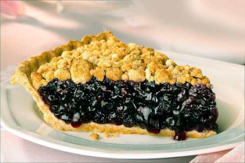 A slice of berry pie with a crumbly topping on a white plate, showcasing a rich, dark filling.