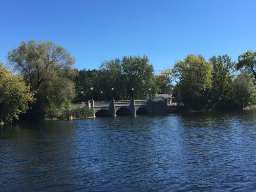 A serene river scene featuring a bridge surrounded by trees under a clear blue sky.