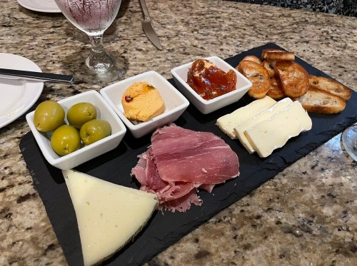 A platter with olives, cheese, cured meat, jam, and crackers on a slate board, set on a granite table.
