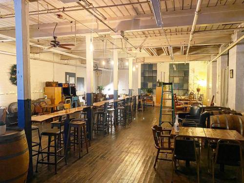 A spacious, empty industrial-style room with wooden floors, tables, and chairs, featuring warm lighting and plants.