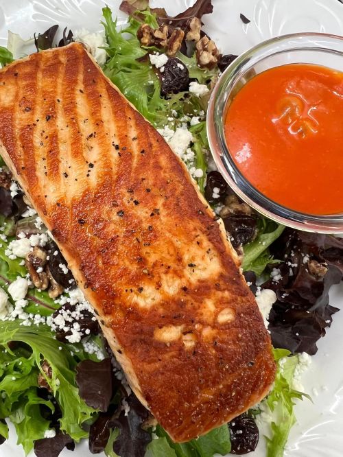 Grilled salmon fillet on a bed of mixed greens with walnuts, feta, and a side of tomato sauce.