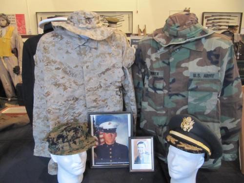 Two military uniforms displayed with hats and framed photos on a table, showcasing service in the Marines and Army.