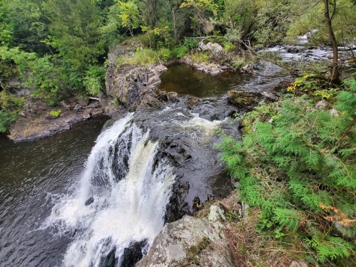 A scenic view of a waterfall cascading over rocky terrain, surrounded by lush greenery and a calm pool below.