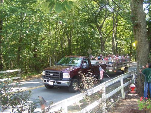 A red pickup truck pulls a trailer with people seated, driving down a tree-lined road.