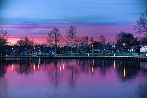 A serene lake at sunset, reflecting vibrant pink and purple skies, with trees and park lights lining the shore.